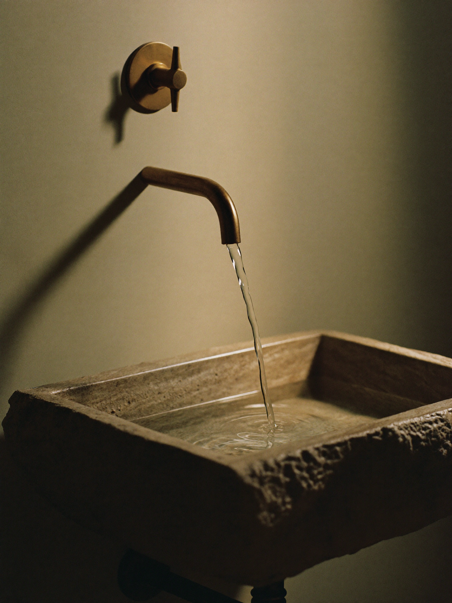 Ruhige Aufnahme eines Messingauslaufs mit langsam tropfendem Wasser in ein honiertes Steinbecken im Abendlicht.